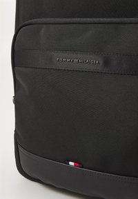Black nylon backpack featuring a smooth texture, branded strap with "TOMMY HILFIGER," and a tri-color flag detail at the bottom.