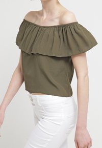 Olivenfarvet off-the-shoulder top med lagdelt ruffle-design, lavet af let stof, parret med hvide højtaljede bukser.