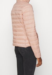 Gepolsterte rosa Jacke mit hohem Kragen, horizontal gestepptem Design und Seitentaschen. Der Stoff wirkt glatt und leicht.