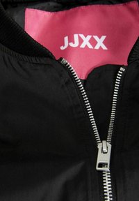 Veste noire avec fermeture éclair argentée partiellement ouverte montrant une étiquette intérieure rose vif avec des lettres blanches "JJXX" sur le col.