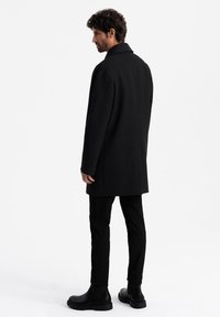 Abrigo negro de mezcla de lana con silueta recta, mangas largas y cuello con muesca, combinado con pantalones negros ajustados y botas negras gruesas.