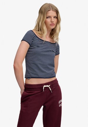 Ung kvinde med blondt hår iført en marineblå og hvidstribet kort t-shirt og bordeaux træningsbukser med hvide snører.