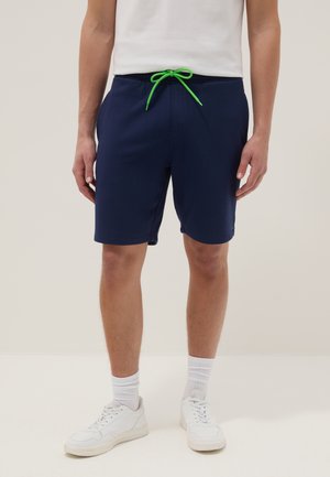Uomo che indossa pantaloncini blu navy con coulisse verde brillante, calzini bianchi, scarpe da ginnastica bianche e una maglietta bianca, in piedi davanti a uno sfondo uniforme.