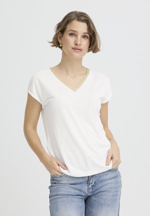 Femeie cu păr scurt și castaniu, purtând cercei aurii, cămașă albă cu decolteu în V și margini aurii, blugi deschis la culoare, cu mâinile în buzunare, fundal neutru.