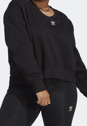 Personne portant un sweat-shirt Adidas noir et un legging avec des logos Adidas blancs, une chaîne en or et des bagues à la main.