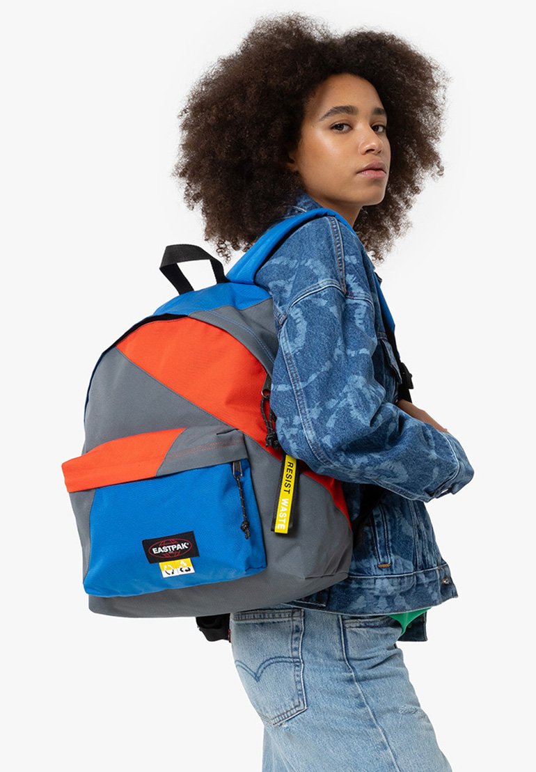 Zaino multicolore con pannelli blu, grigi e arancioni, una tasca anteriore e un'etichetta gialla, realizzato in tessuto resistente.