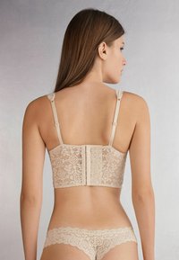 Beige lace bra with intricate floral patterns, wide back band, adjustable straps, and hook closure. Matching lace briefs with scalloped edges.