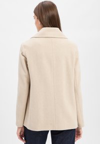 Beige Wollmischung Mantel mit Reverskragen, Fischgrätmuster, einzelner Naht hinten und Fronttaschen, getragen über dunklen Jeans.