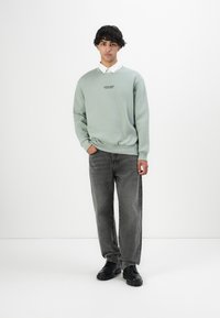 Jonge man staat in grijze jeans, zwarte laarzen, lichtgroene sweatshirt over wit colbert, tegen een effen witte achtergrond.