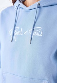 Felpa leggera azzurra realizzata in morbido tessuto. Presenta la scritta ricamata in bianco "Project x Paris" e occhielli in tono argento sul cappuccio.
