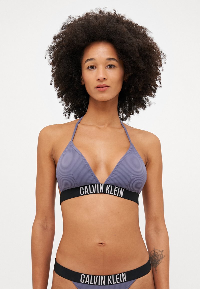 Fialový bikiny top s ribovaným vzorem a trojúhelníkovými košíčky, s černým logem na pásku. Odpovídající bikiny dolní část s podobným designem. Hladký povrch.