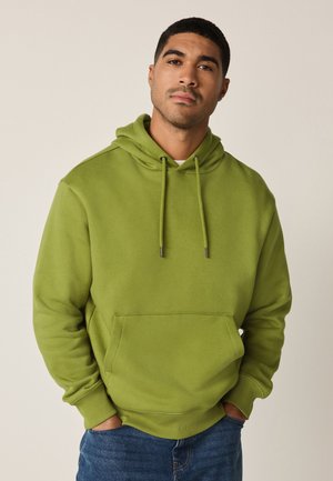 Man wearing a green hooded sweatshirt with drawstrings and front pocket, standing with hands in pockets against a plain background.