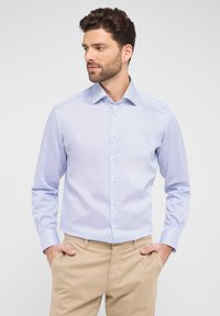 Helles blaues Hemd mit einem kleinen, dunklen Punktmuster. Mit Knopfleiste, spitzem Kragen und langen Ärmeln. Getragen mit beigen Hosen.