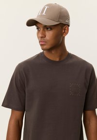 Καφέ κοντομάνικη μπλούζα με τετράγωνο λογότυπο στο στήθος και μπεζ καπέλο μπέιζμπολ με λευκές γραμματοσειρές και καμπύλη γείσο.