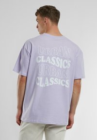 Levandulové bavlněné tričko s velkým bílým nápisem "URBAN CLASSICS" v hravém písmu vytištěným na zadní straně, s krátkými rukávy a volným střihem.