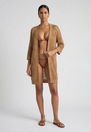 Kobieta ubrana w brązowe bikini i pasujący otwarty sweter z dzianiny oraz sandały, stojąca na jednolitym jasnym tle.