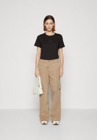 Svart kortermet skjorte, beige cargo-bukser med sidelommer, hvit buet veske og teksturerte hvite sneakers, mot en ensfarget bakgrunn.