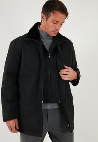 Černý kabát z vlněné směsi s vysokým límcem a dvojitým předním zipem, vybavený bočními kapsami a měkkou vnitřní podšívkou.