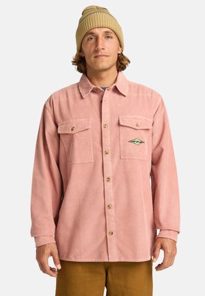 Camisa de pana en rosa claro con cierre de botones, dos bolsillos en el pecho y un detalle de logo bordado, combinada con un gorro de punto beige.