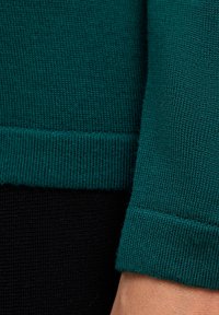 Tessuto di maglione in teal con orlo a coste, che mostra dettagli ravvicinati della texture e del motivo di cucitura su uno sfondo nero.