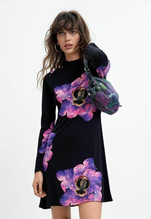 Mujer con vestido negro con grandes estampados de flores rosas y moradas, llevando un bolso texturizado multicolor en el hombro.