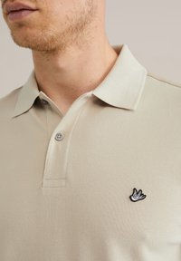 Tricou polo bej dintr-un material texturat, cu două nasturi și un logo brodat subtil al unei păsări pe partea stângă a pieptului. Design cu guler.