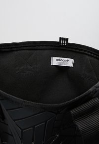 Svart Adidasväska med slät yta, dragkedja ovanpå och foder i vävt tyg. Har en etikett inuti med märkesdetaljer och design med tre ränder.