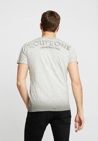 Camiseta de color gris claro, de manga corta, con un diseño desvanecido en la parte posterior. El texto está impreso en gris oscuro y rojo. Tejido suave, corte ajustado.