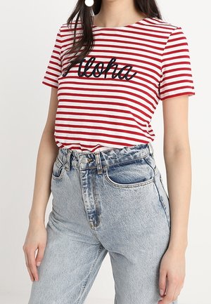 Frau trägt rot-weiß gestreiftes T-Shirt mit "Aloha" Text und hoch taillierte hellblaue Jeans aus Denim vor weißem Hintergrund.