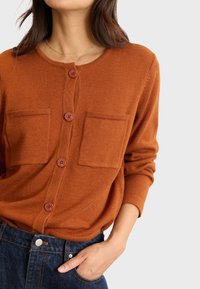 Cardigan lavorato a maglia di color ruggine, con colletto rotondo, cinque bottoni e due tasche frontali, abbinato a jeans di denim scuri.