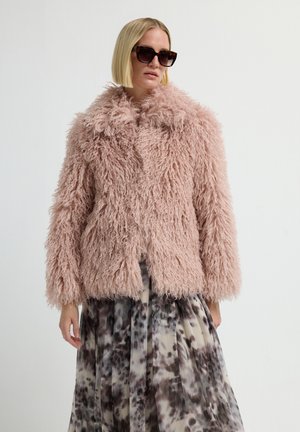 Woman wearing a fluffy pink fur jacket, patterned skirt, and large dark sunglasses against a plain white background.
