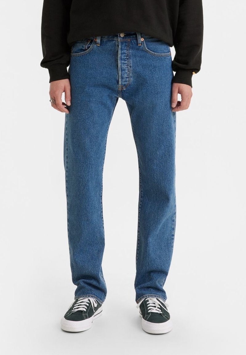 Personne portant un jean bleu droit, un sweat-shirt noir et des baskets basses noires à lacets blancs, debout sur un fond blanc.