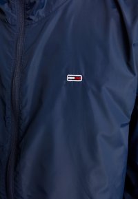 Granatowa kurtka przeciwwiatrowa z gładkiego nylonu, z zamkiem błyskawicznym i małym haftowanym logo w czerwonych i białych akcentach.