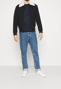 Chaqueta bomber negra con cuello de borrego blanco, combinada con un suéter azul marino con lunares y unos vaqueros de denim azul claro. Unas zapatillas blancas completan el look.