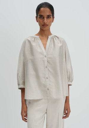 Femme aux cheveux foncés coiffés en chignon bas, portant un chemisier ample en satin beige clair avec des boutons et un pantalon large assorti, sur un fond uni.