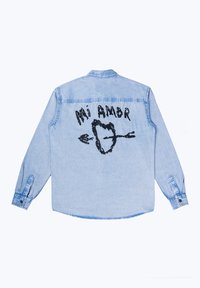 Világoskék farmering hosszú ujjakkal, fekete grafikai nyomattal, amelyen szív és nyilak láthatók, rajta a "Mi Amor" felirattal a hátsó részén.