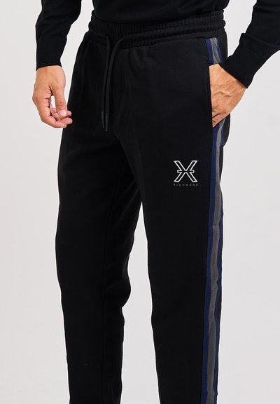 Hombre con pantalones de chándal negros con bandas laterales en azul y gris, mano en el bolsillo, y logo blanco de "Richmond" en el muslo.