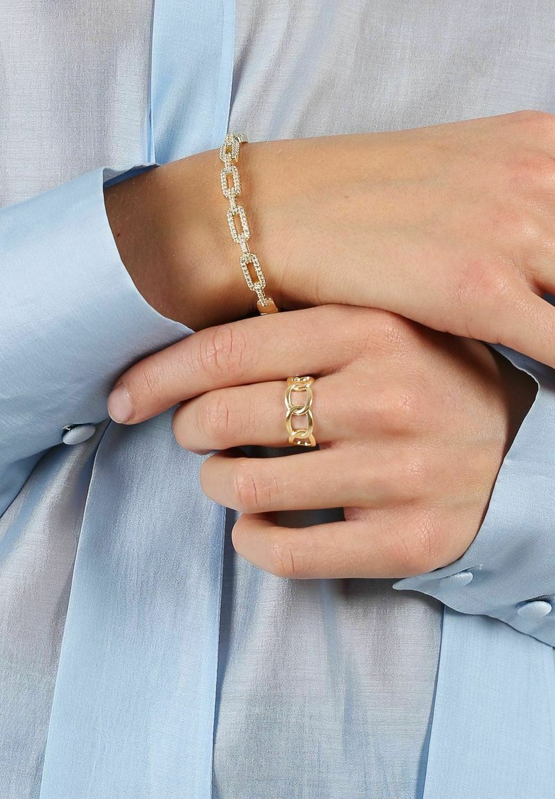 Goldkette mit strukturierten Gliedern und einem goldenen Ring mit ineinandergreifenden Formen, getragen an einer Hand auf einem hellblauen Hemd.