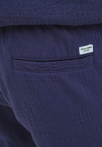 Tissu texturé bleu marine avec une petite étiquette de patch indiquant "JACK & JONES EST. 1990" et un détail de poche arrière.