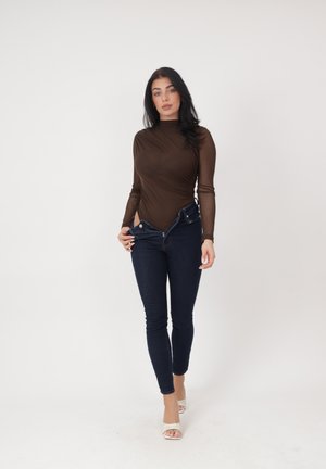 Kvinde iført en brun langærmet top og mørkeblå jeans med åbne knapper, gående fremad mod en ensfarvet hvid baggrund.