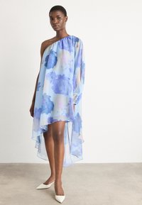 Asymmetrisk blå klänning i genomskinligt tyg med blommönster, en lång ärm och en svävande fåll. Matchad med vita spetsiga skor.
