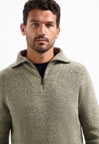 Man met donker krullend haar en baard, gekleed in een lichtgrijze trui met halfrits over een zwart shirt, tegen een witte achtergrond.