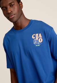 Blauwe T-shirt met korte mouwen van katoen, met een roze en groene grafische print van "Ciao" met een reis-thema op de linkerkant van de borst.