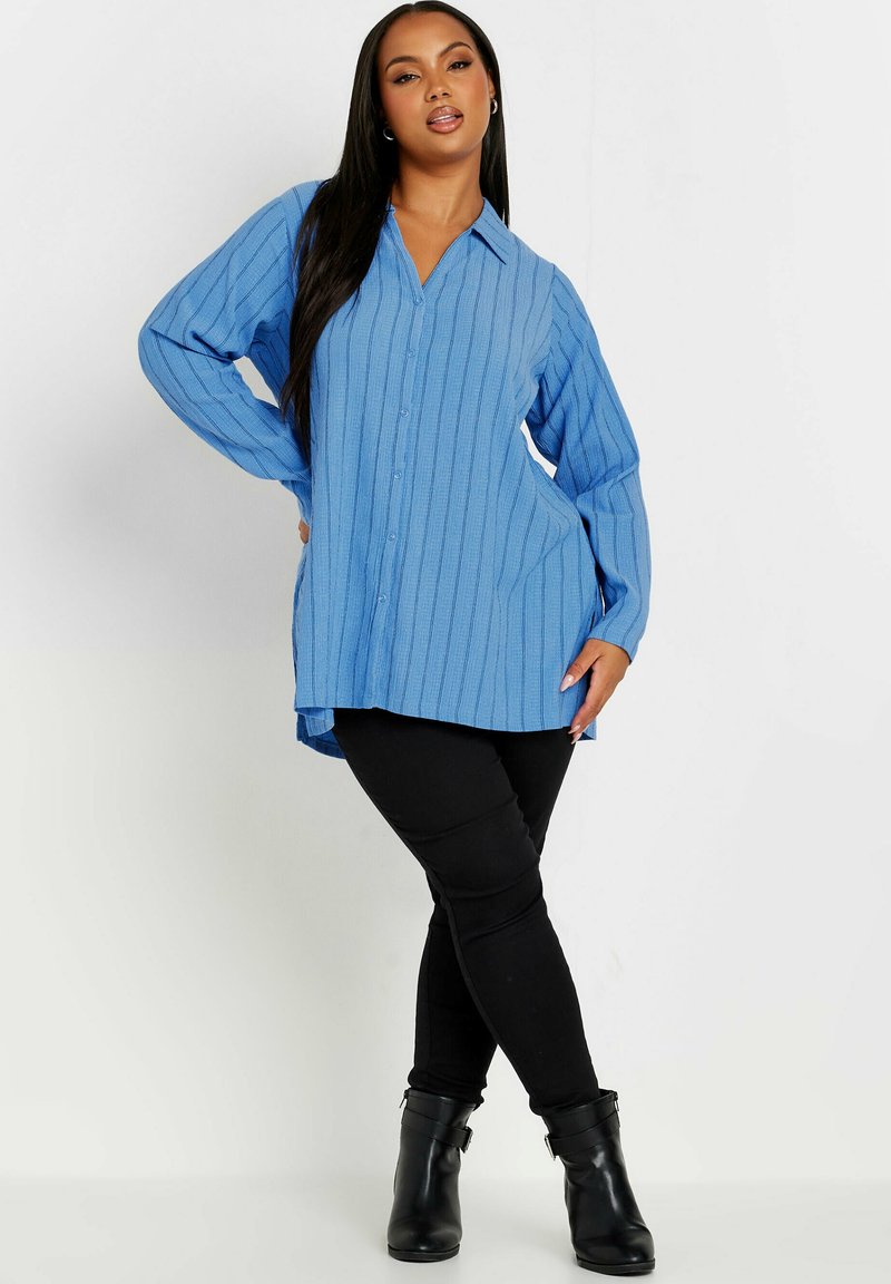 Camicia button-up azzurro chiaro con texture a righe verticali, maniche lunghe e colletto, abbinata a leggings neri e stivali alla caviglia.