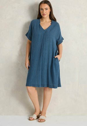 Kobieta stojąca w pomieszczeniu, ubrana w luźną niebieską sukienkę do kolan z krótkimi rękawami oraz beżowe plecione płaskie sandały, z jedną ręką w kieszeni.