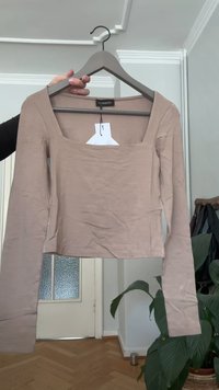 Beige langärmliges Cropped-Top mit quadratischem Ausschnitt auf einem grauen Kleiderbügel, von einer Hand gehalten in einem Raum mit Holzboden und Pflanzen.