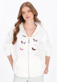 Hanorac alb cu fermoar, decorat cu motive colorate brodate cu câini, o fundă și text. Materialul este moale, cu tiv și manșete striate.