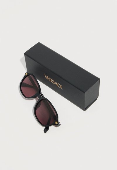 Μαύρα γυαλιά ηλίου Versace με σκοτεινούς φακούς δίπλα σε μαύρη θήκη με το χρυσό λογότυπο της Versace, σε απλό ανοιχτό φόντο.