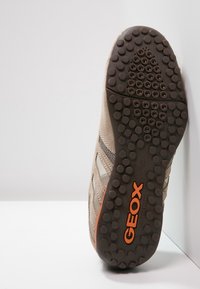 Sula av beige mockasinsneaker med runda svarta grepp, orange GEOX-logotyp och nätstoffdetaljer, placerad upprätt mot vit vägg.