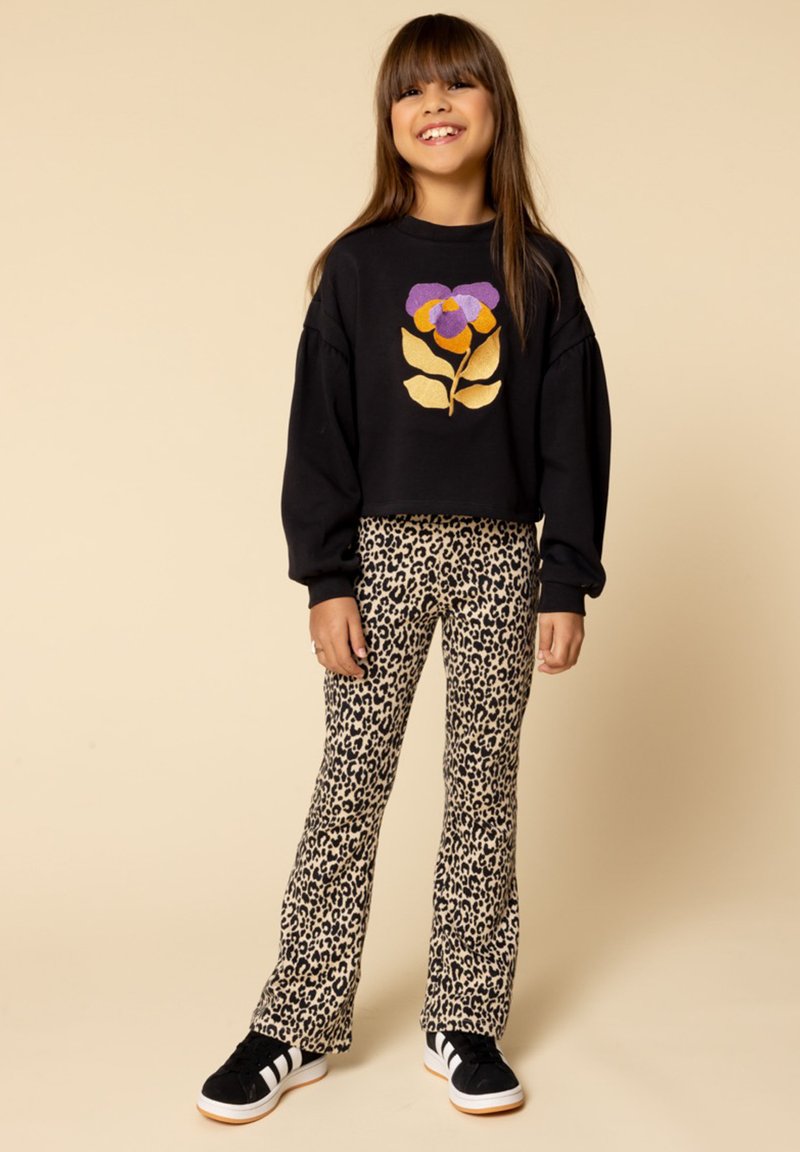 Zwarte cropped trui met een kleurrijk bloemenontwerp, gecombineerd met beige flared broek met luipaardprint en zwarte sneakers met witte strepen.